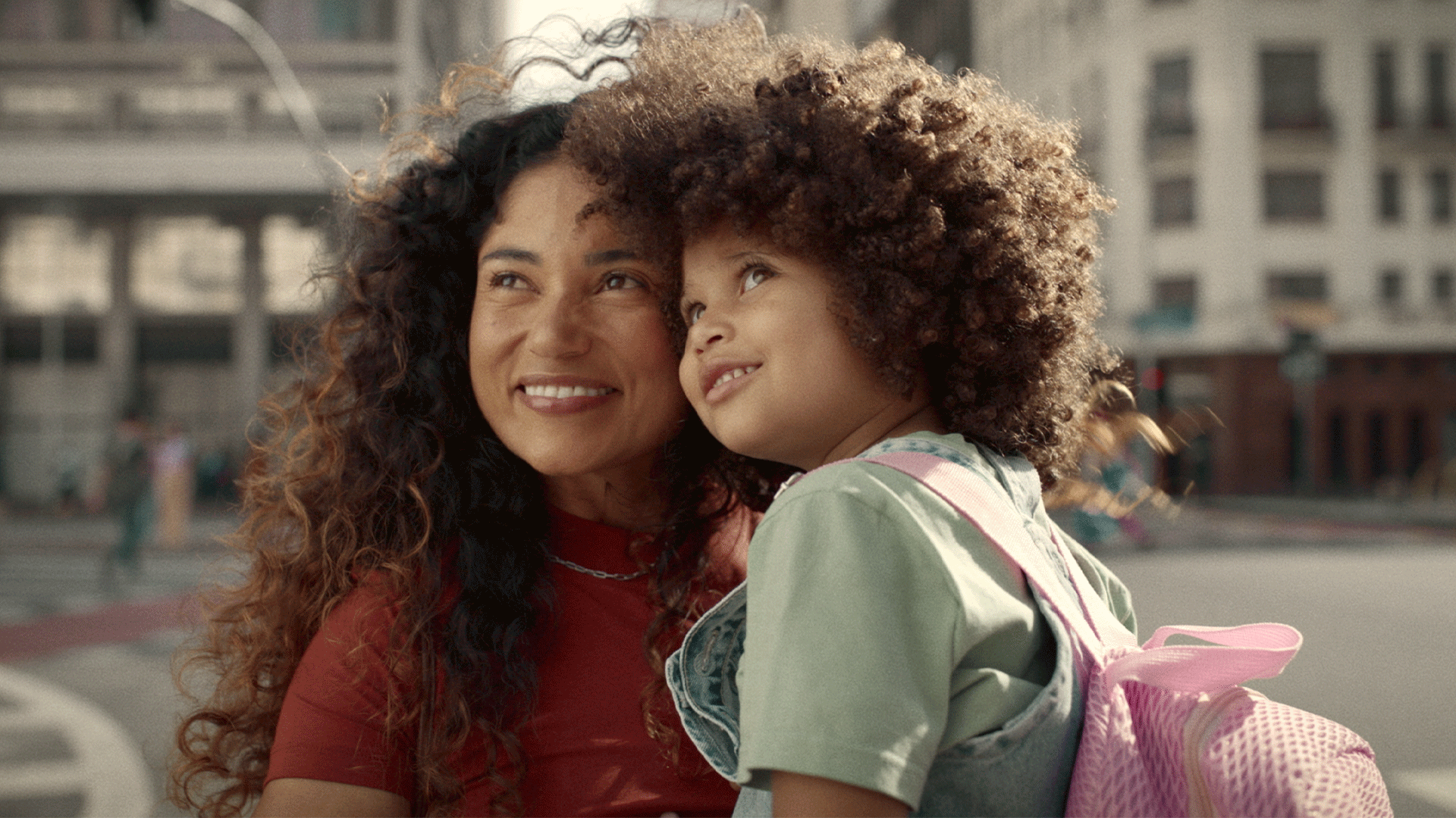 A woman holding a child with a pink backpack, with a city street in the background. They are both smiling.