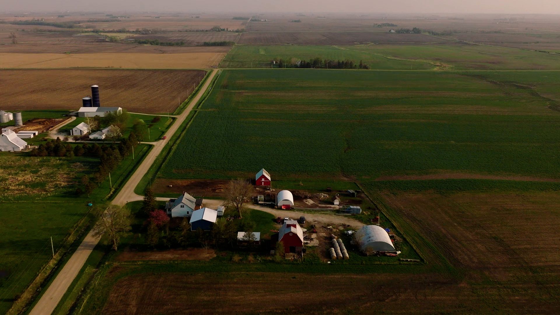 Aerial view of some rural farmland with several buildings including barns, surrounded by vast expanses of green and brown agricultural fields.