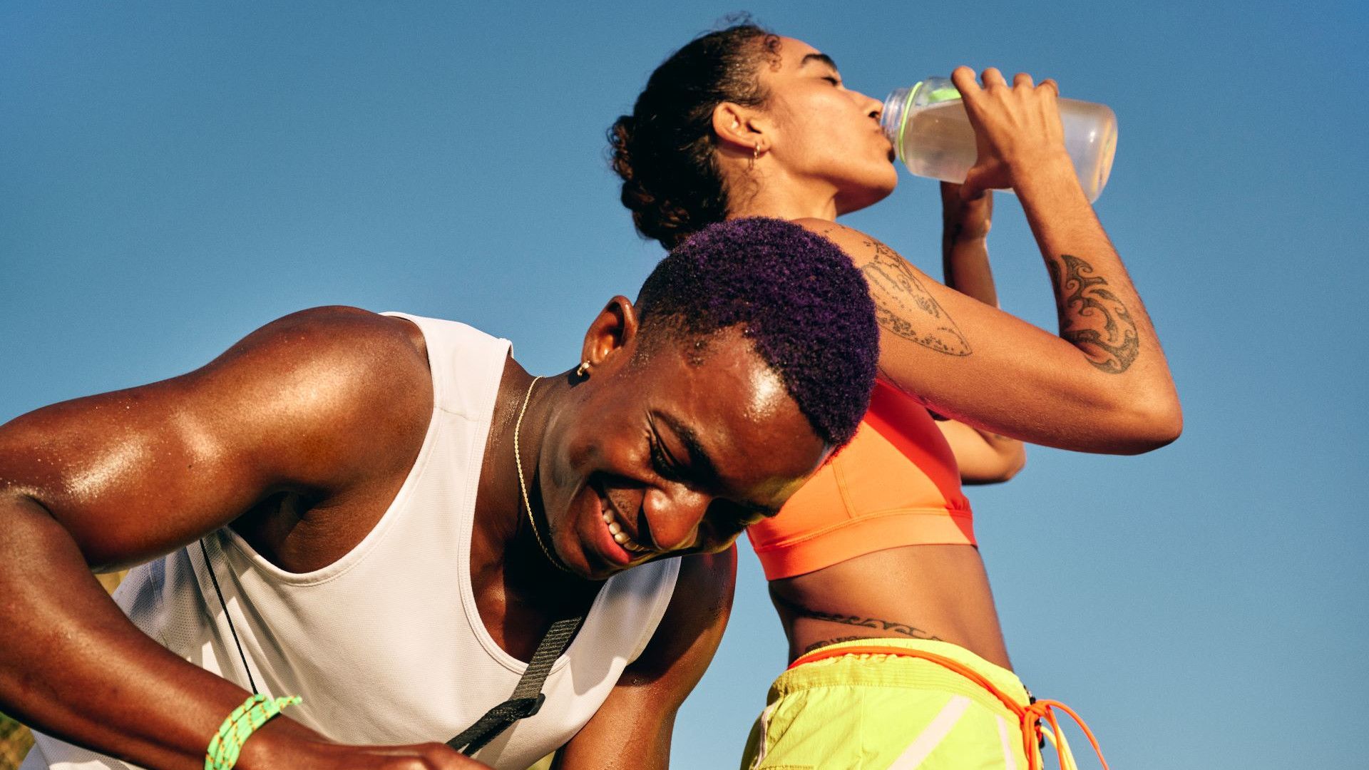 Man smiles as the woman beside him drinks from a bottle of Liquid I.V. and water during a pause in their outdoor activity.
