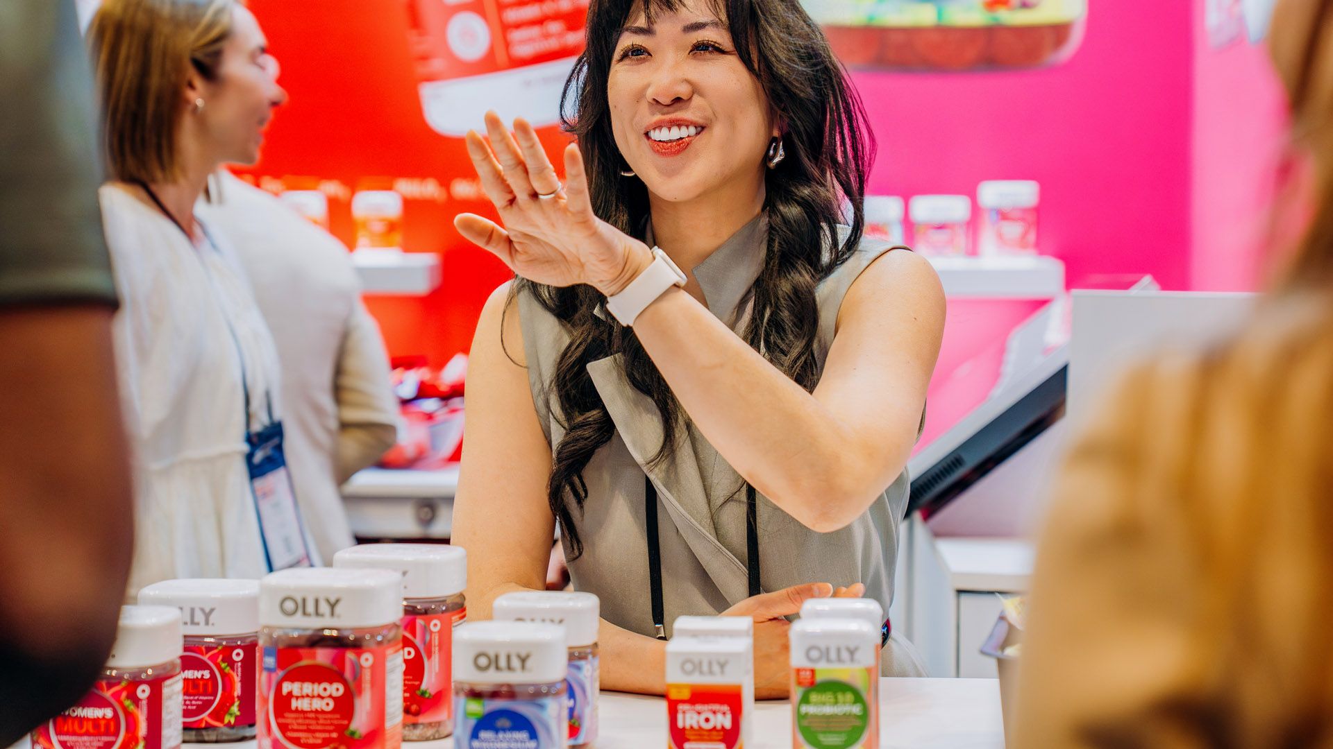 Smiling woman, standing behind a table upon which sits a range of Olly products. the woman is talking to a person who is out almost out of frame.