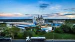 Industrial site with tanks, buildings, and a tanker truck on a road, set against a blue sky with clouds and greenery.