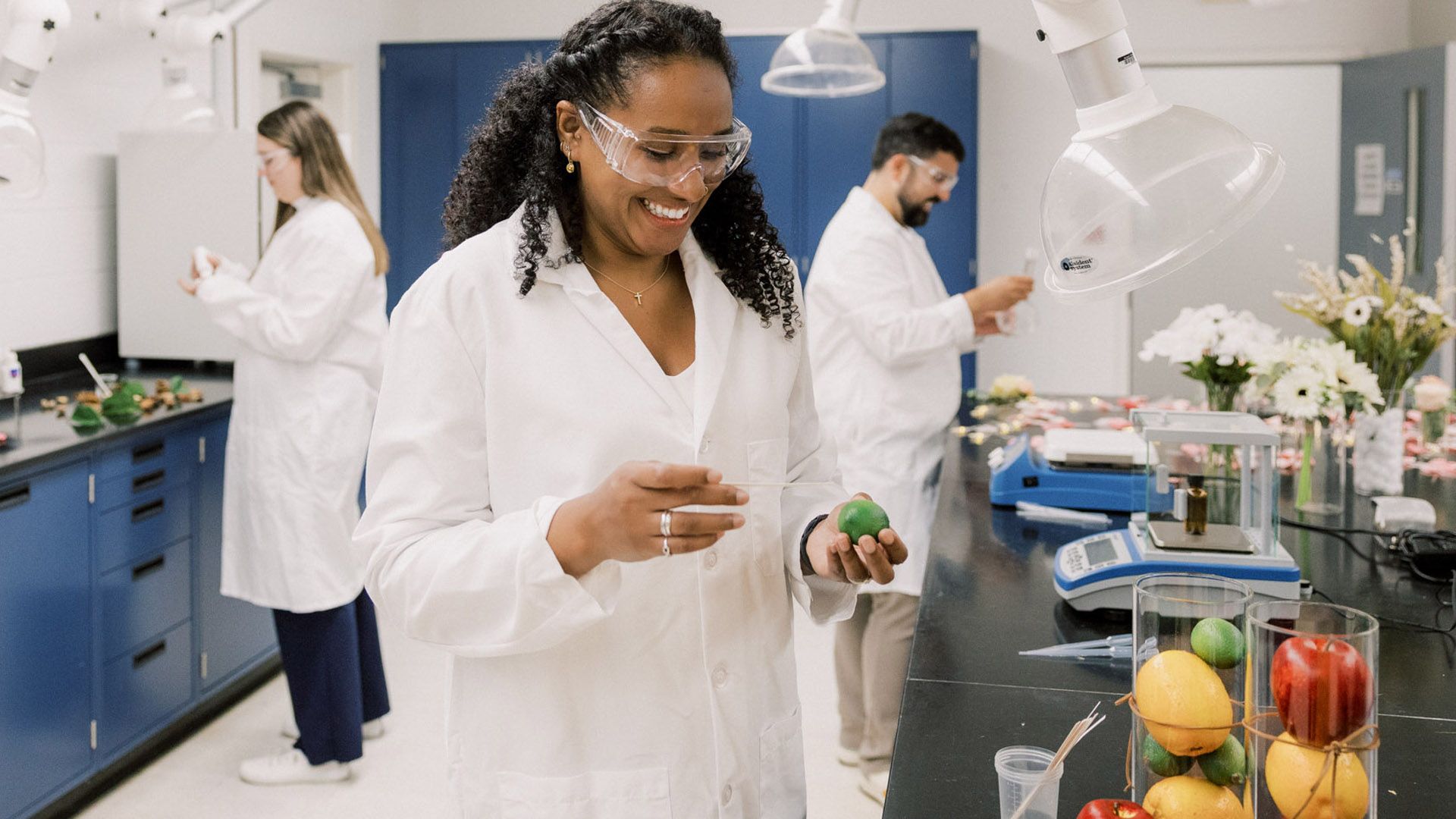 Three people in a fragrance lab conducting experiments with various flowers and items of fruit.