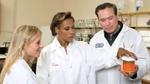 Three people in white lab coats standing in a science lab look closely at a small jar full of a chemical substance.