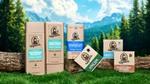 Dr. Squatch grooming products – including shampoo, conditioner, deodorant, and soaps – arranged on logs and grass in a natural outdoor setting with trees, mountains, and a blue sky in the background.