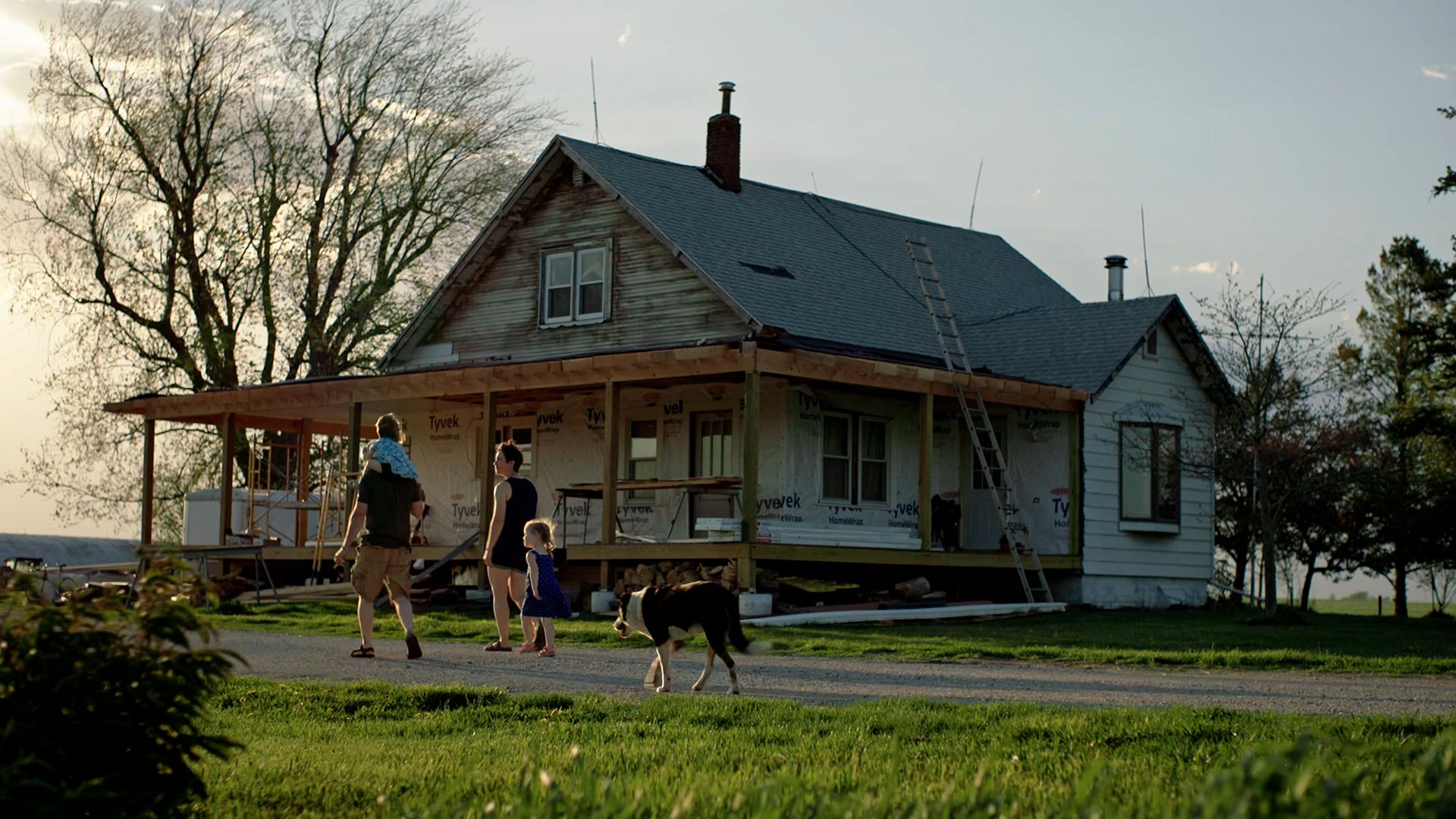 A man with a child on his shoulders, a woman, a small girl and a dog walk up to a farm house