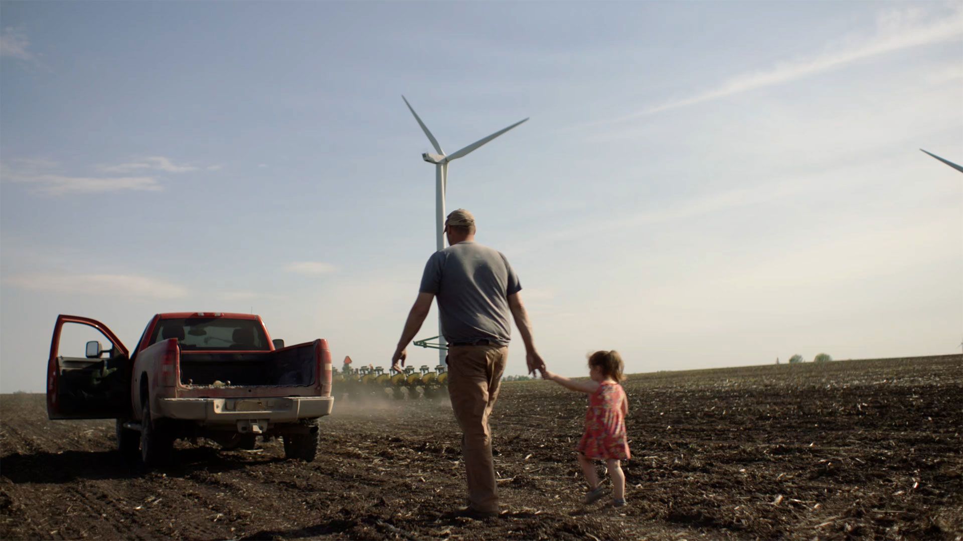 A man in a baseball cap holds the hand of a little girl while walking across a ploughed field to a pick-up truck.