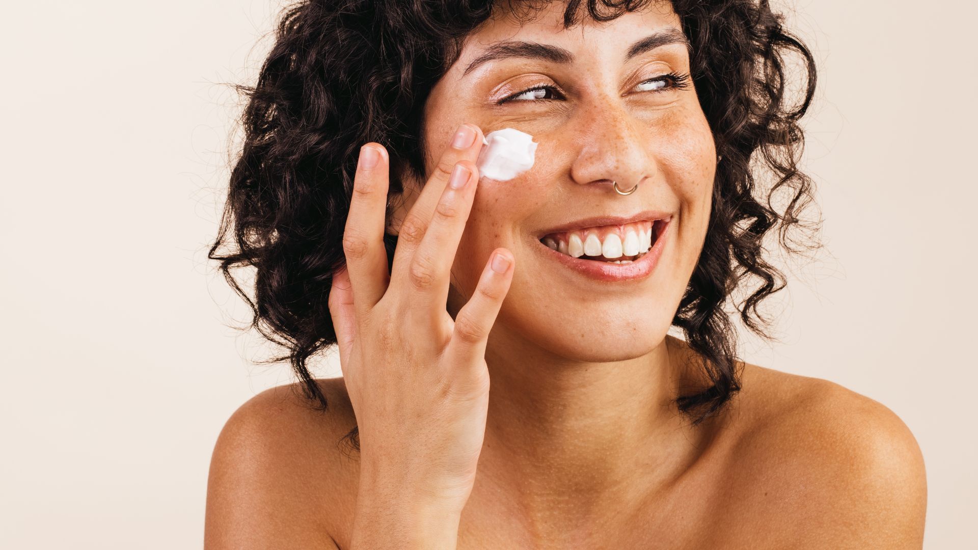 A woman with short dark curly hair smiles while applying a small dab of white face cream to their cheek with their fingers